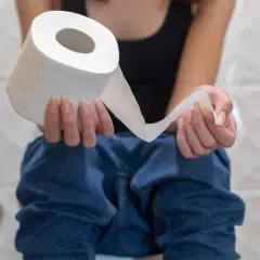 Osoba siedząca na toalecie trzymająca rolkę papieru toaletowego, sugerująca problemy z biegunką.