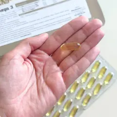 Gold Omega 3 na co pomaga? Jedna kapsułka w dłoni, obok opakowania i blistra. Wspiera serce i mózg.