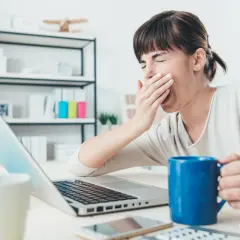 Kobieta zmagająca się z hipersomnią ziewa przy laptopie, trzymając kubek kawy.