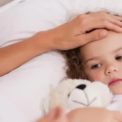 Matka czule dotyka czoła chorej córki, która leży w łóżku z misiem. Objawy zapalenia oskrzeli u dzieci mogą być niepokojące.