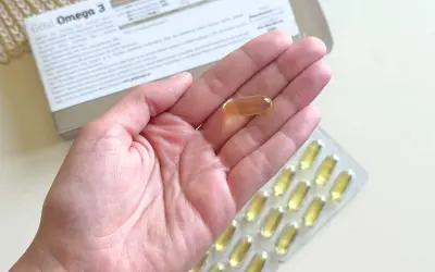 Gold Omega 3 na co pomaga? Jedna kapsułka w dłoni, obok opakowania i blistra. Wspiera serce i mózg.