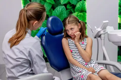 Dziewczynka z niepokojem czeka na usunięcie zęba, podczas gdy dentystka w rękawiczkach przygotowuje się do zabiegu.