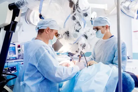 Chirurdzy w maseczkach i fartuchach operacyjnych pracują przy użyciu mikroskopu.