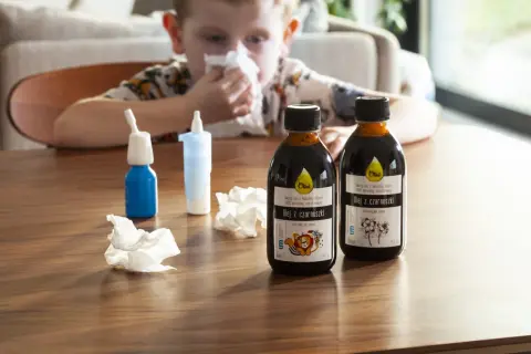 Chłopiec z katarem sięga po olej z czarnuszki, szukając domowych sposobów na uczulenie skórne. Na stole stoją butelki oleju i spray do nosa.