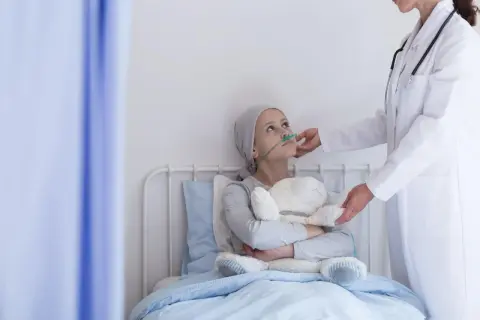 Dziewczynka z rakiem serca w szpitalnym łóżku, z tlenem i pluszowym misiem, otrzymuje wsparcie od lekarki.