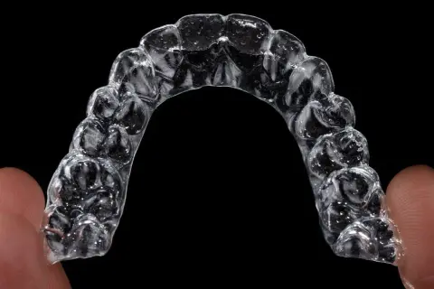 Przezroczysty aparat Invisalign, trzymany palcami, z widocznymi wypustkami dopasowanymi do zębów.