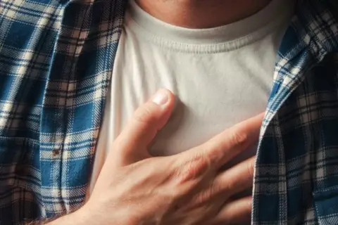 Dłoń na piersi, odczuwając niepokojący ucisk w klatce piersiowej. Mężczyzna w koszuli w kratę.