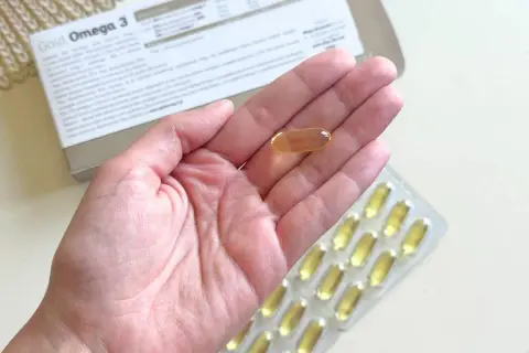 Gold Omega 3 na co pomaga? Jedna kapsułka w dłoni, obok opakowania i blistra. Wspiera serce i mózg.