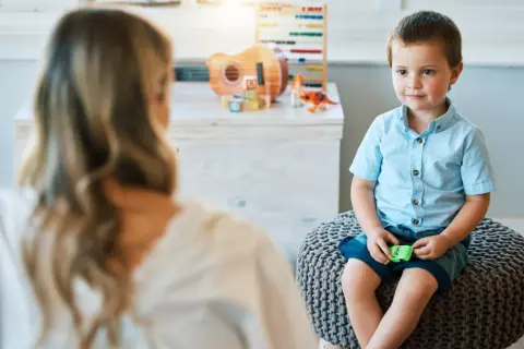 Chłopiec w niebieskiej koszuli siedzi na pufie, rozmawiając z psychologiem.