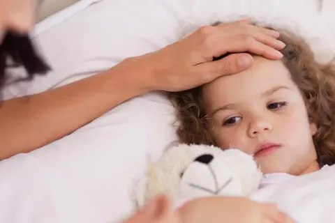 Matka czule dotyka czoła chorej córki, która leży w łóżku z misiem. Objawy zapalenia oskrzeli u dzieci mogą być niepokojące.