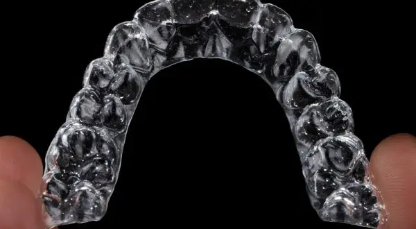 Przezroczysty aparat Invisalign, trzymany palcami, z widocznymi wypustkami dopasowanymi do zębów.