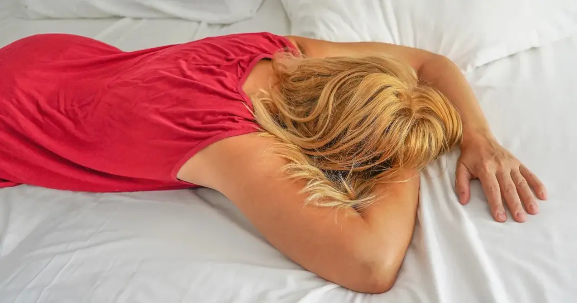 Kobieta w czerwonej sukience leży na białej pościeli, przytulając głowę do ramion. Jej blond włosy opadają na twarz, symbolizując walkę z depresją lękową.