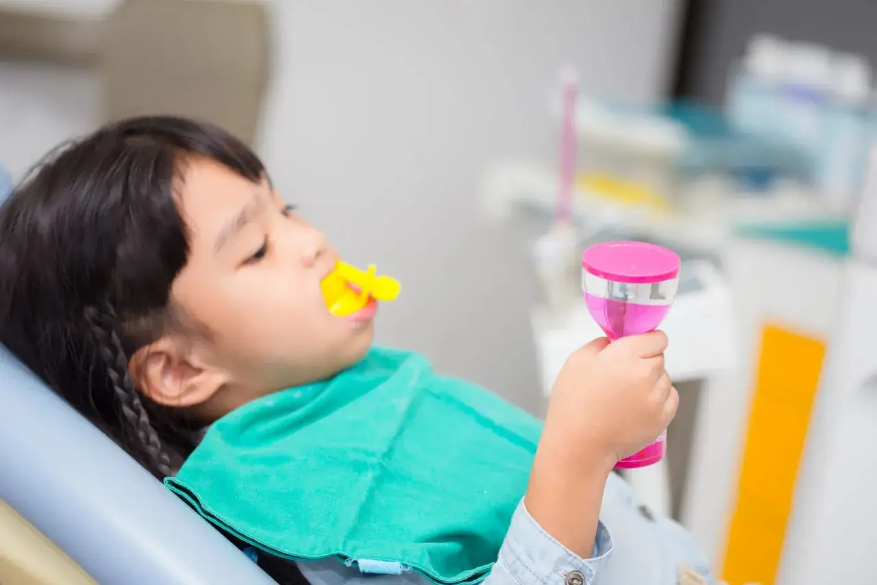 Dziecko leży na fotelu dentystycznym, ma założony żółty retraktor i zielony śliniak. W ręku trzyma różowy kubeczek, być może po zabiegu fluoryzacji.