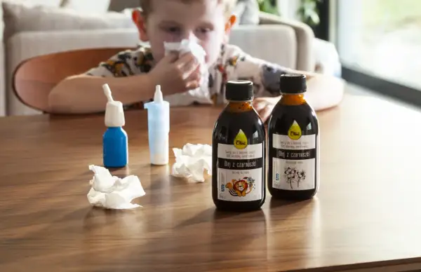 Chłopiec z katarem sięga po olej z czarnuszki, szukając domowych sposobów na uczulenie skórne. Na stole stoją butelki oleju i spray do nosa.